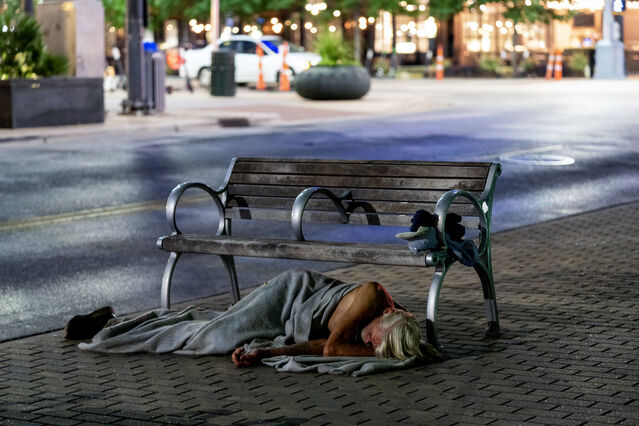 Photograph of a public bench with armrests at either end and in the middle, therefore meaning no-one could lie on it. In front on the bench, on the pavement, a homeless person sleeps rough.